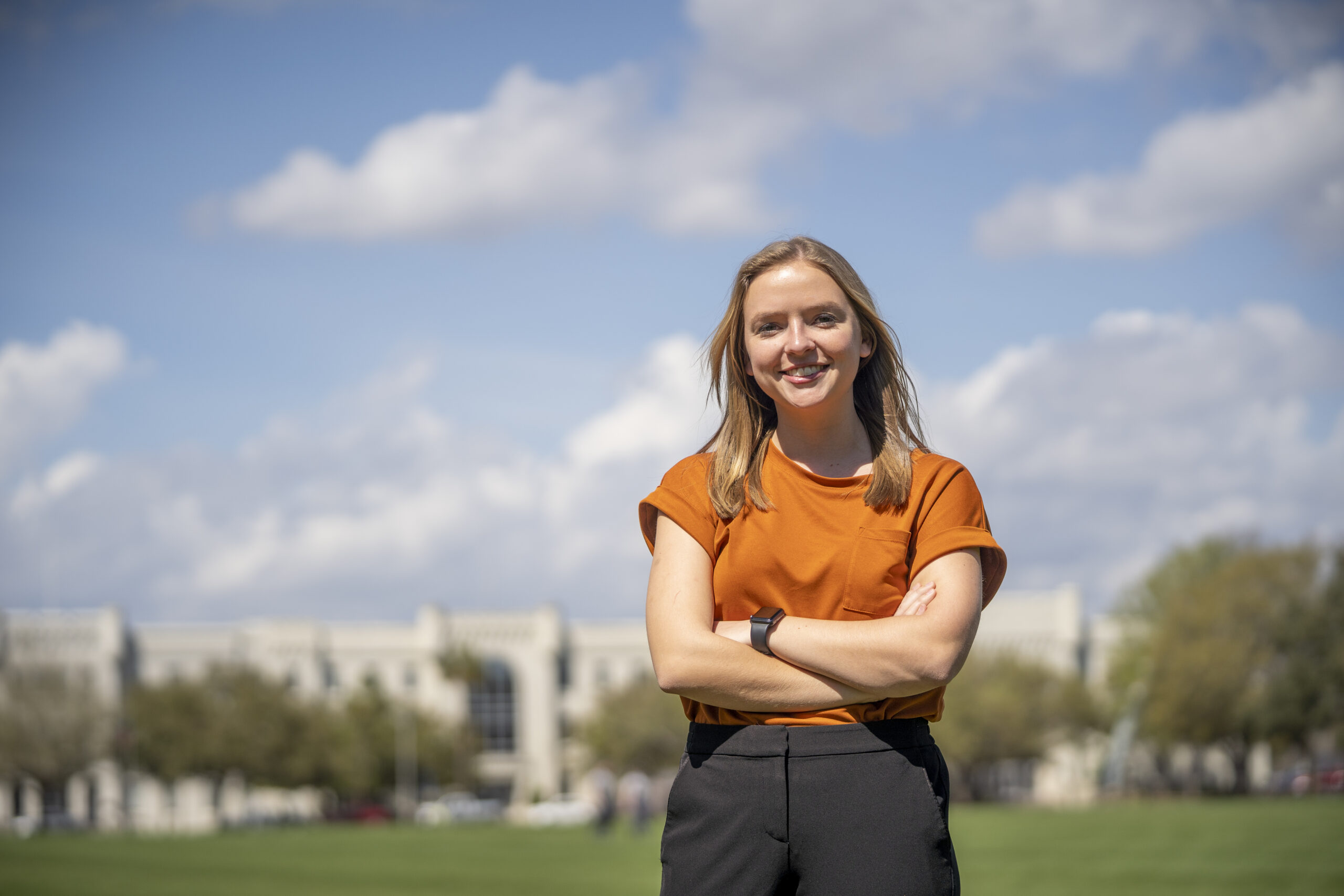 student stands with arms crossed on a campus lawn under a blue sky.
