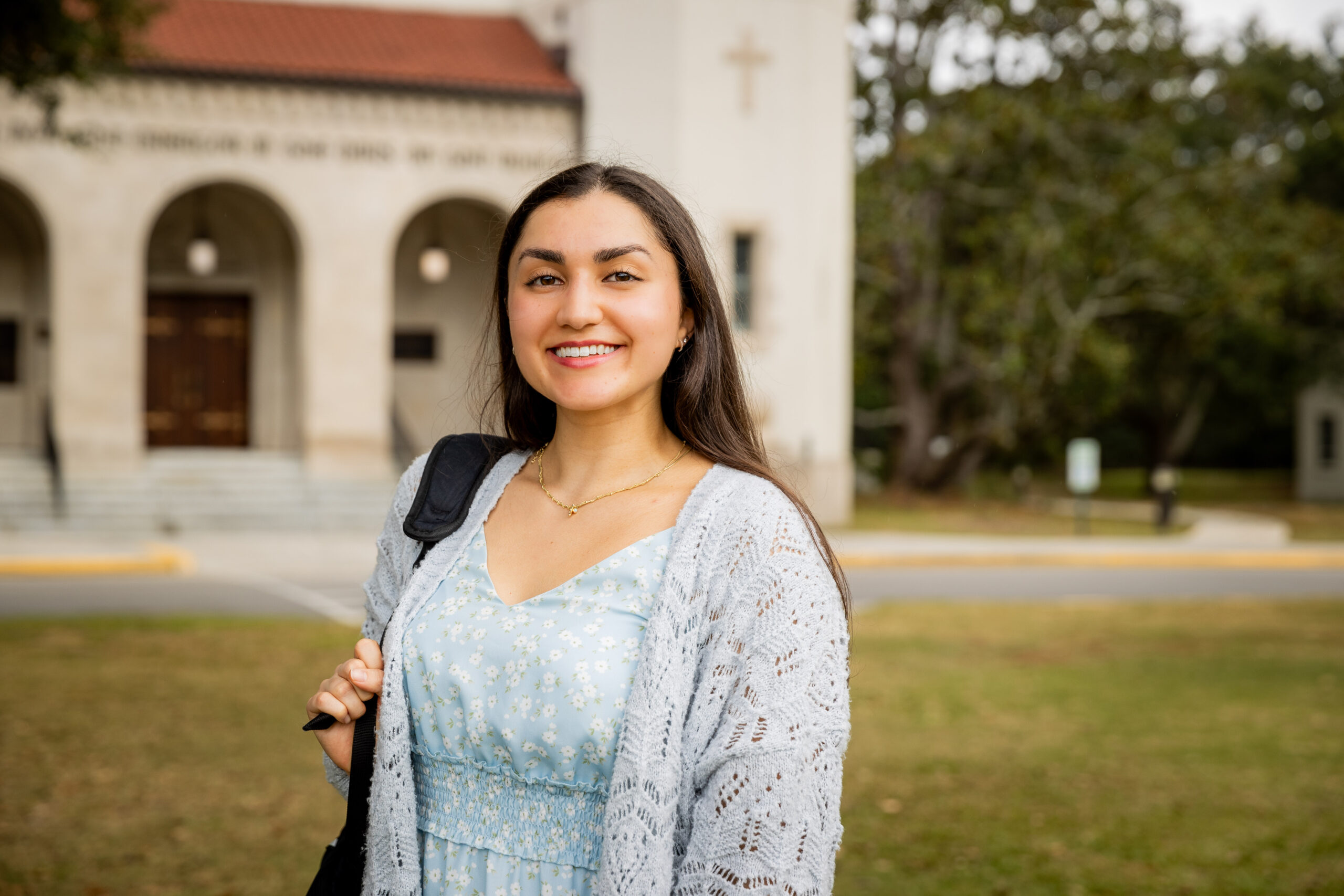 student carrying a backpack smiles outside a campus building.