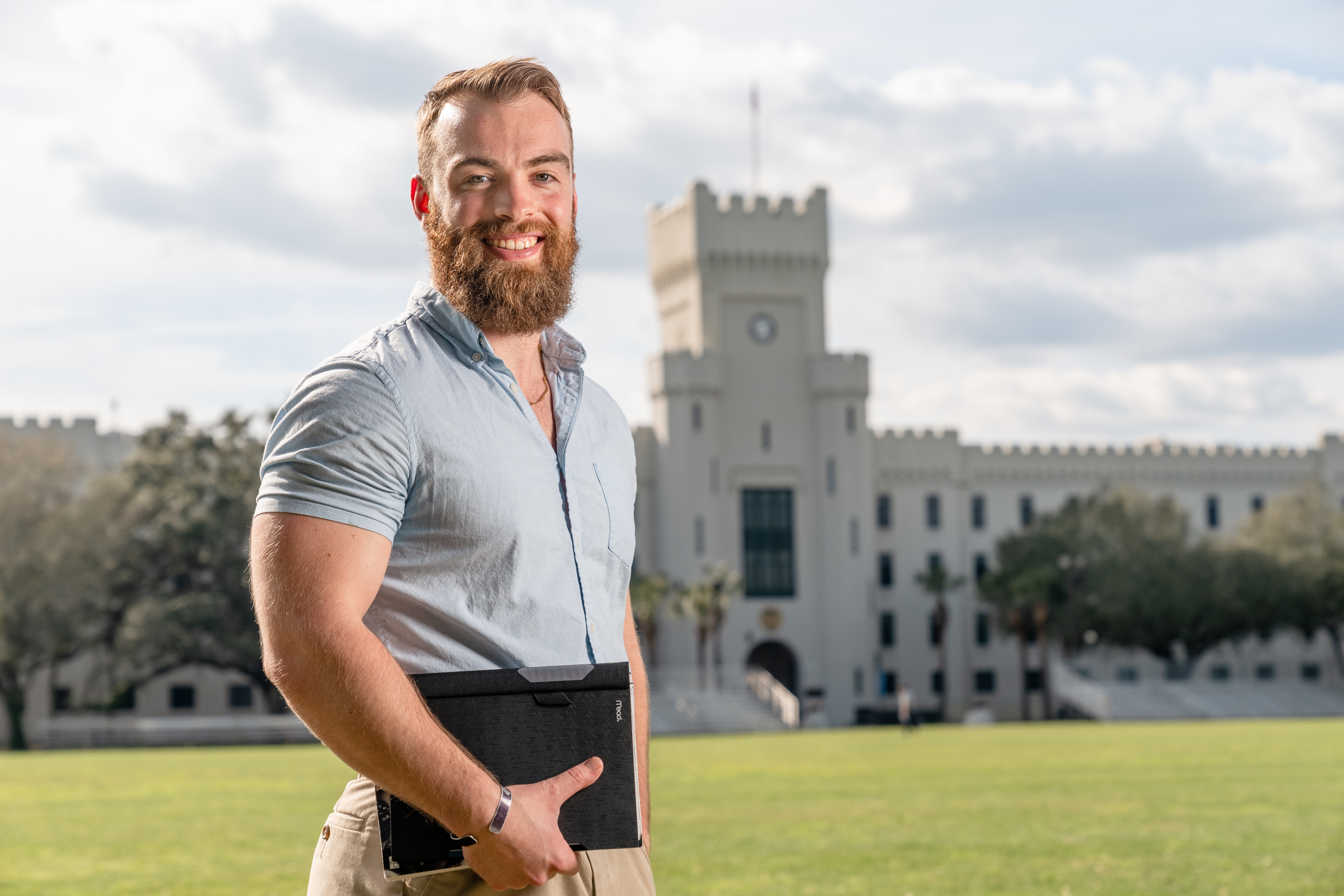 Bearded student holding a portfolio stands on a campus field with a Citadel building in the background.