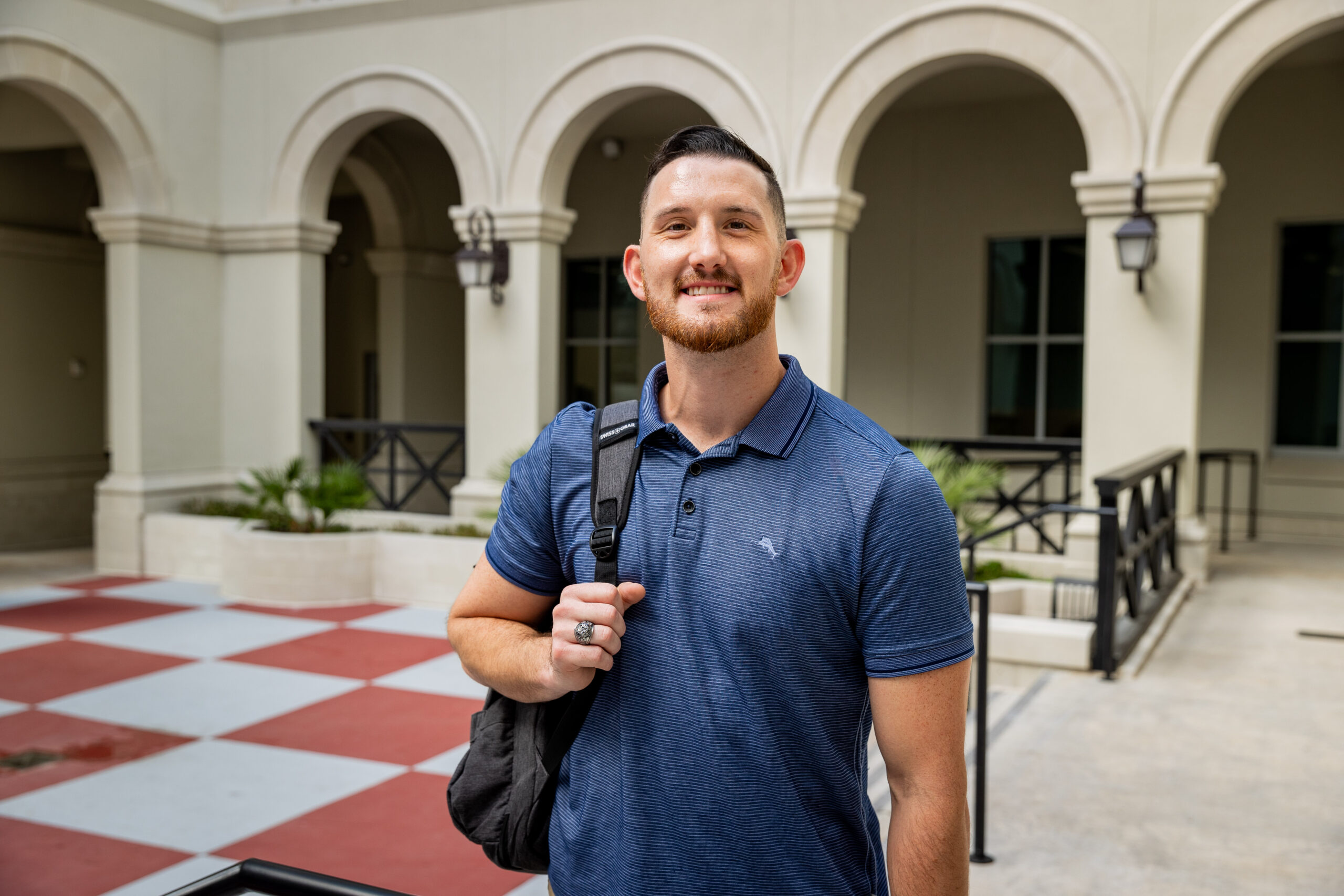 student carrying a backpack smiles in a campus courtyard.