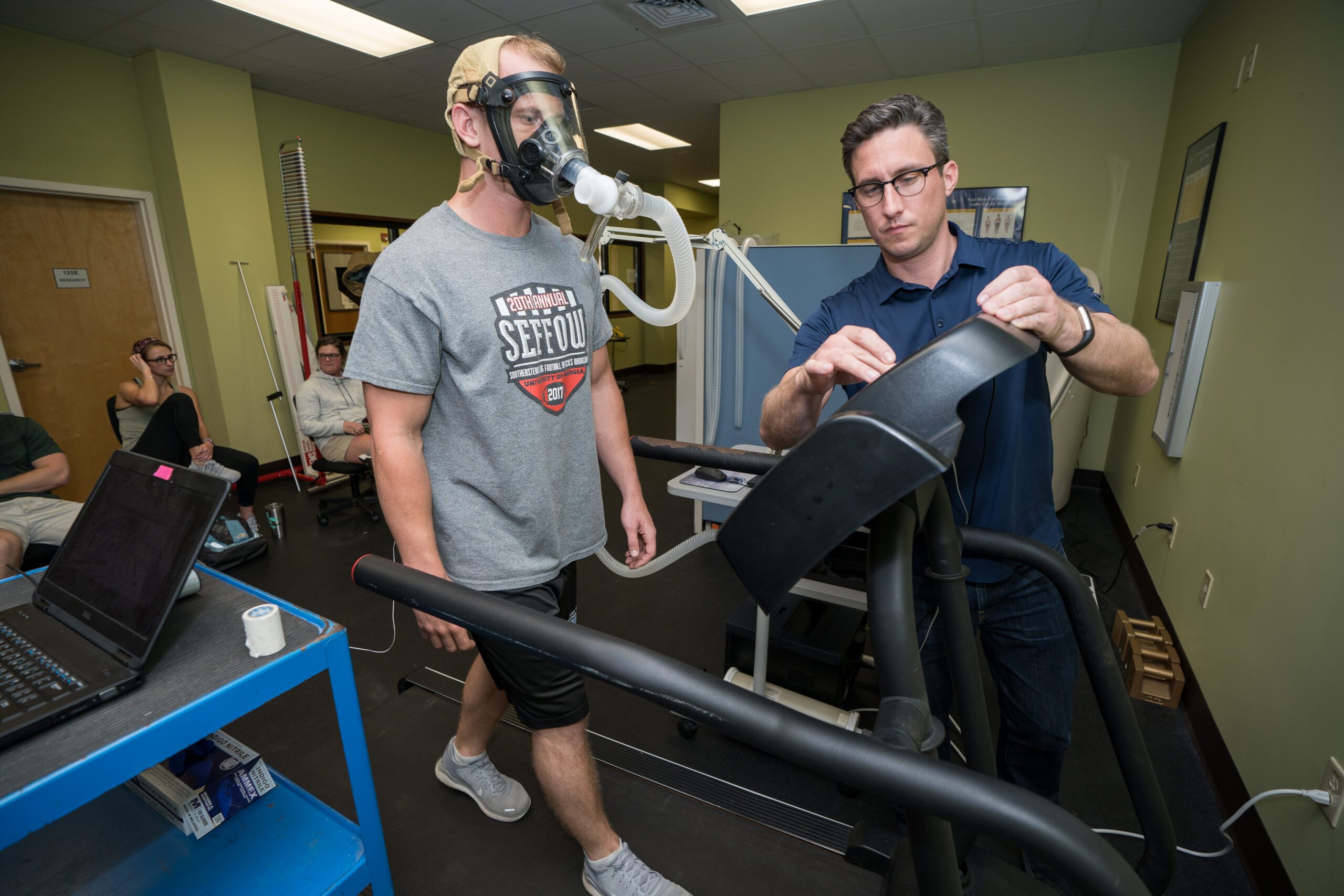 Exercise science student adjusts equipment while a participant wearing a respiratory mask walks on a treadmill during an exercise test.