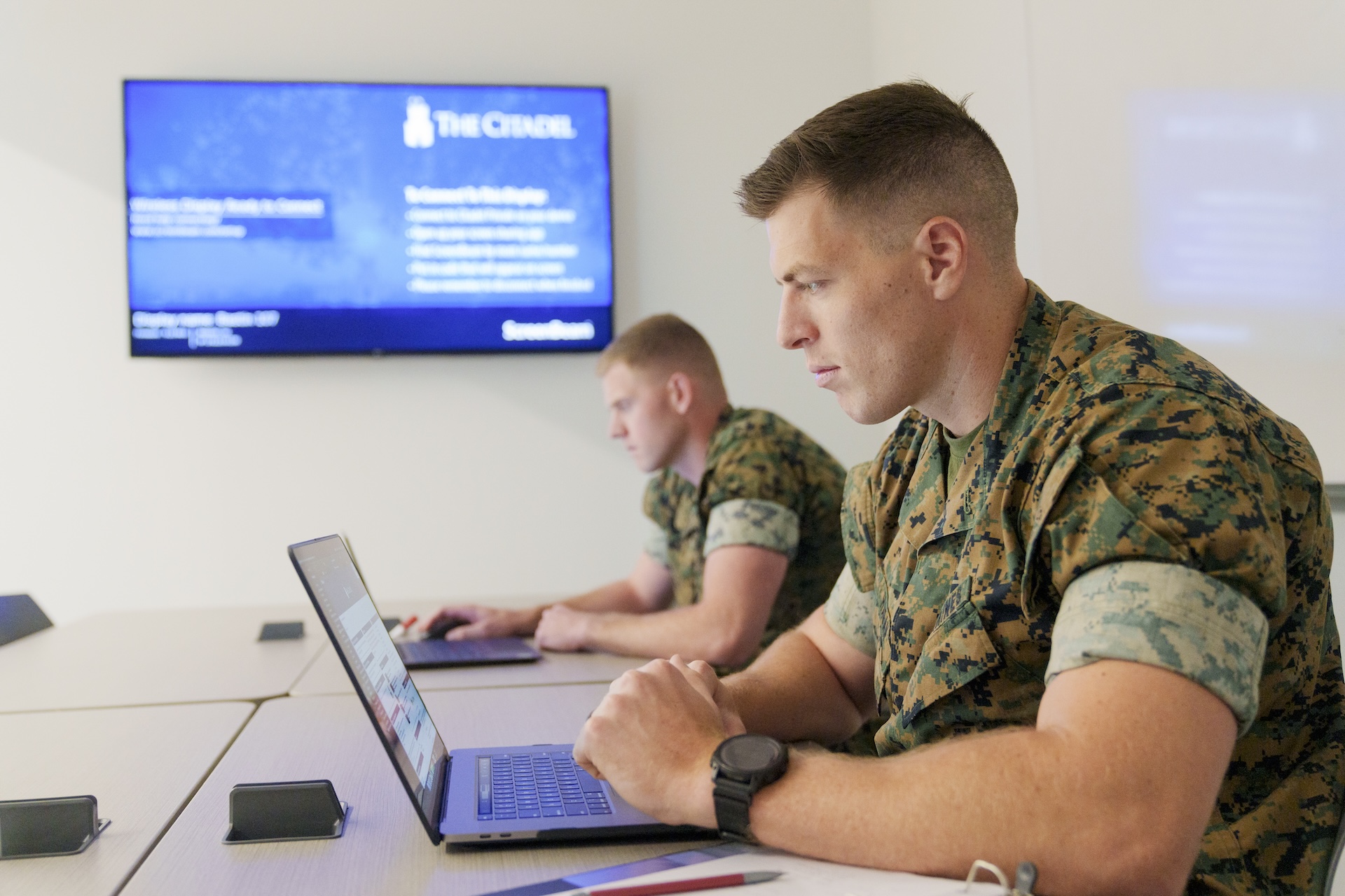 Two active duty students in camouflage uniforms work on laptops in a classroom.