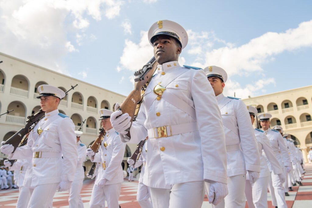 Cadets in white dress uniforms stand in formation with rifles on The Citadel’s checkerboard courtyard.