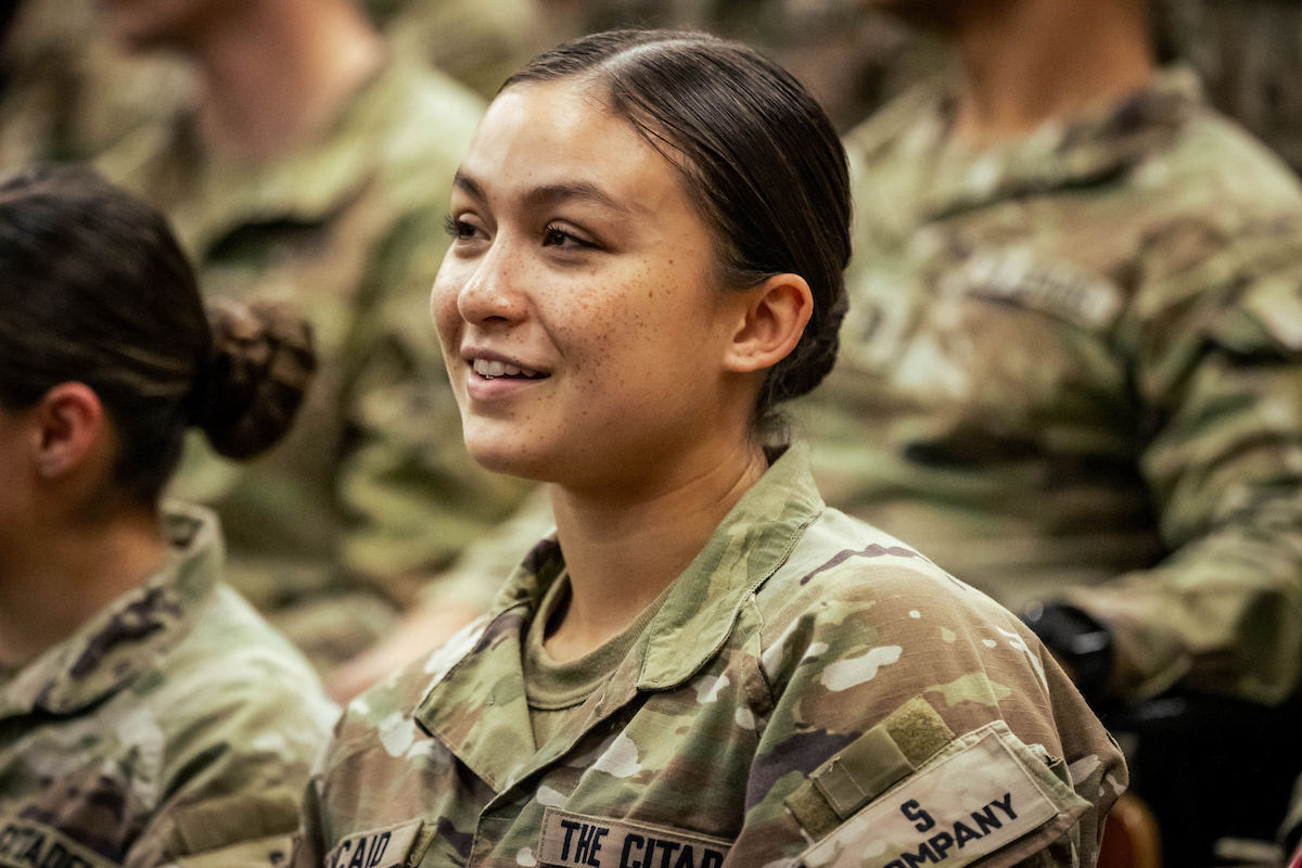 Citadel cadet in a military uniform listens during an event with other cadets seated nearby.
