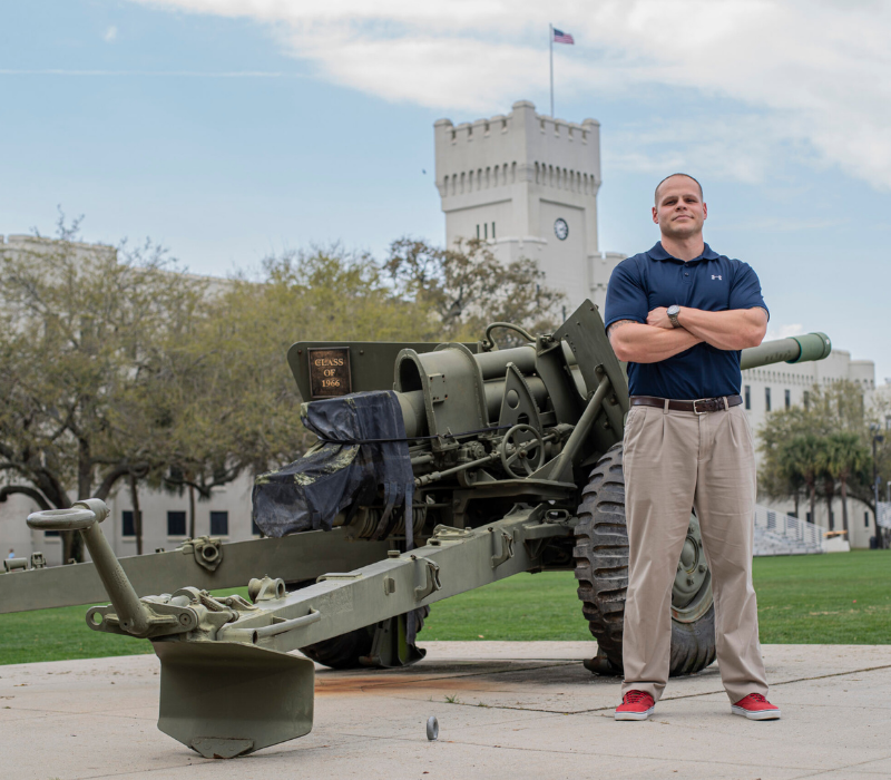 Graduate student stands with his arms crossed beside a field artillery piece on The Citadel campus.