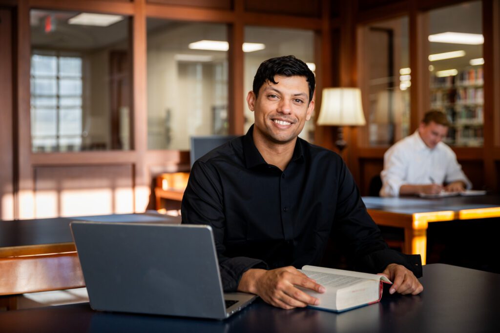 Man in a black shirt studies with an open book and laptop in Daniel Library.