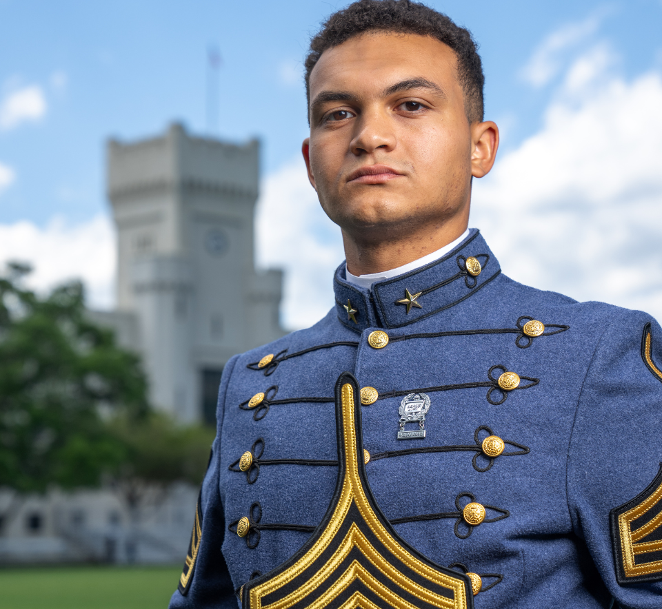 Citadel cadet in dress uniform holds a large chevron insignia while posing outdoors.